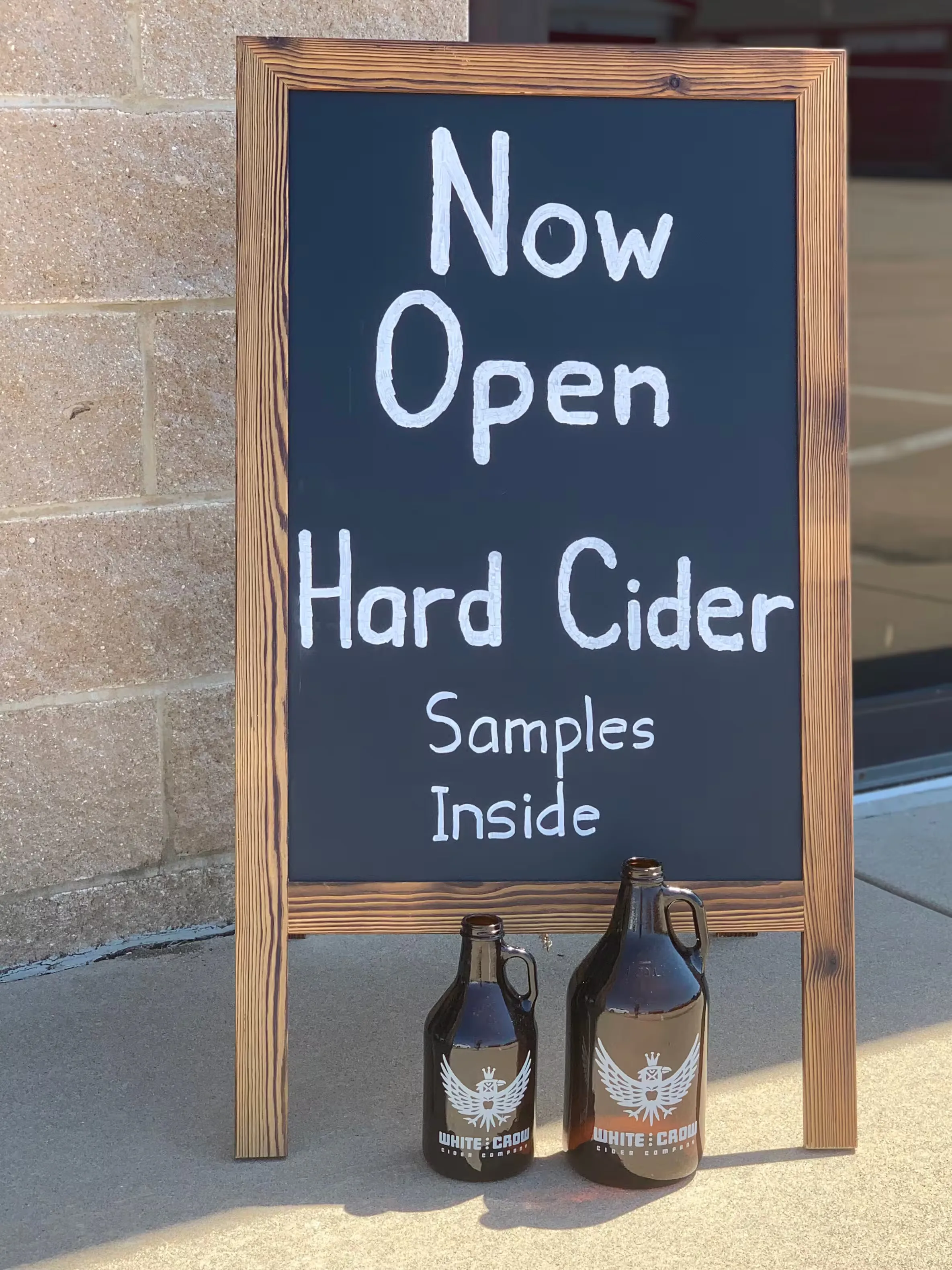 Wooden chalkboard sign reading ‘Now open – hard cider samples inside.’
